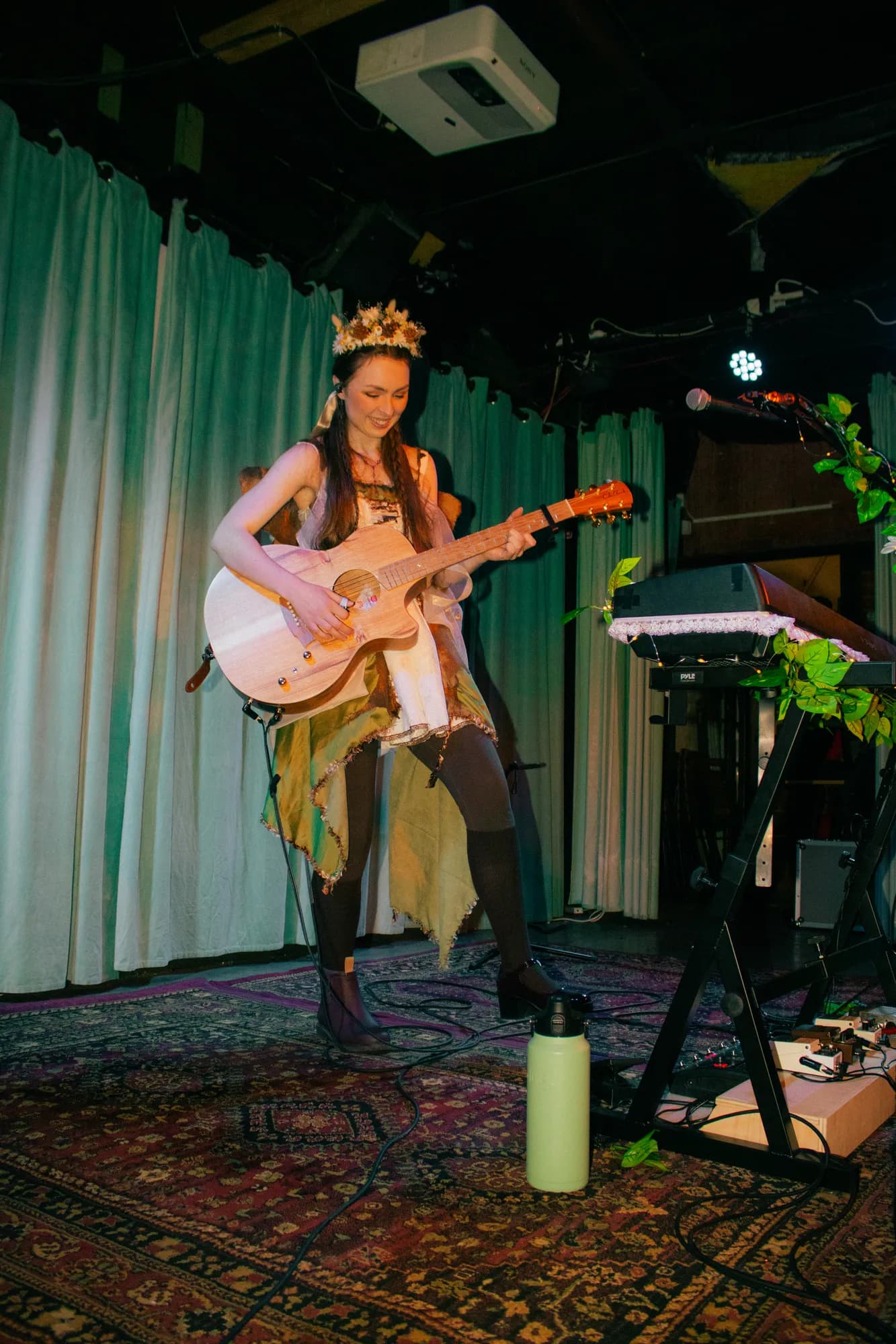 Solly Fern in a flower crown playing acoustic guitar on a patterned rug, her vine-dressed keyboard beside her, at The Jade, July 2026