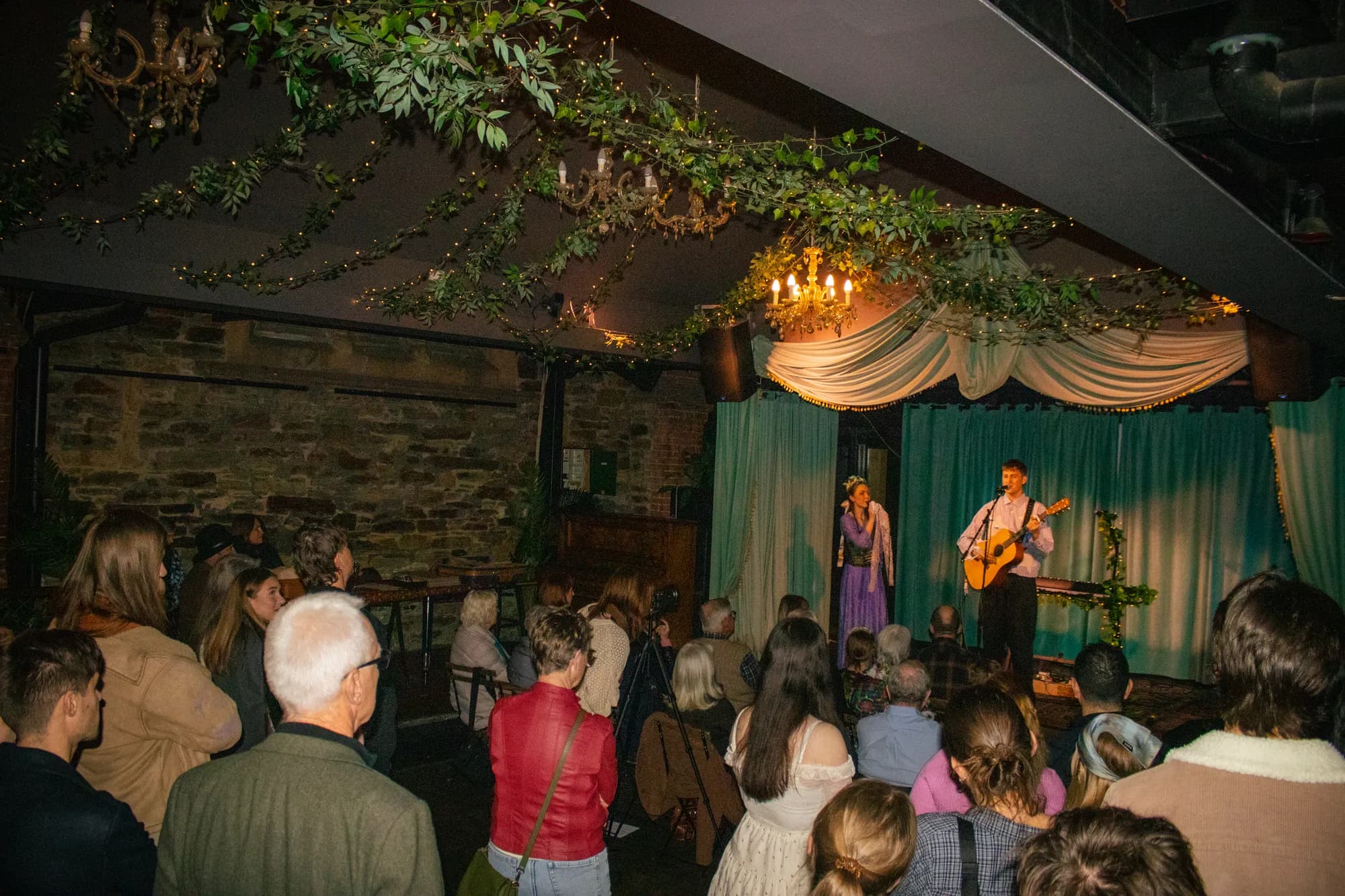 A full room at The Jade watching Solly Fern and a guitarist on a stage dressed with vines, drapes and a chandelier, at the Into Halves EP launch, July 2026