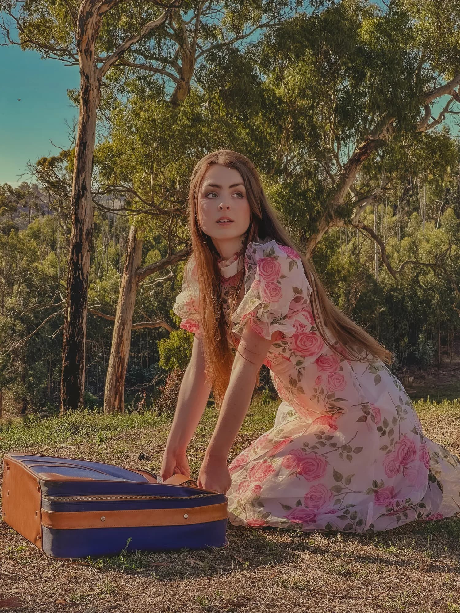Solly Fern in a rose print dress kneeling beside an old suitcase in the bush